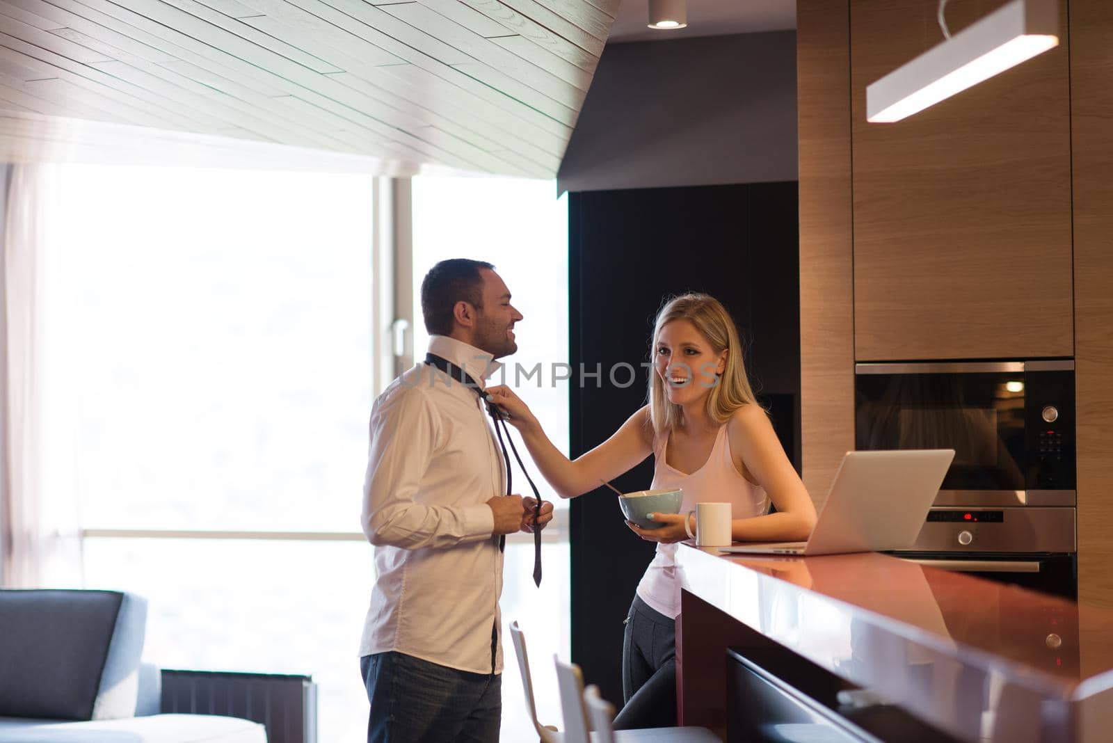 A young couple is preparing for the job and using a laptop. The man drinks coffee while the woman eats breakfast at luxury home together, looking at screen, smiling.