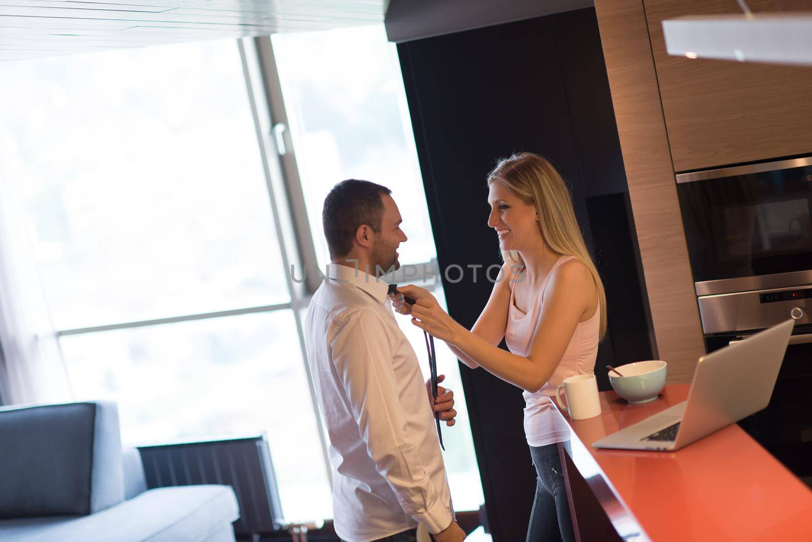 A young couple is preparing for the job and using a laptop. The man drinks coffee while the woman eats breakfast at luxury home together, looking at screen, smiling.