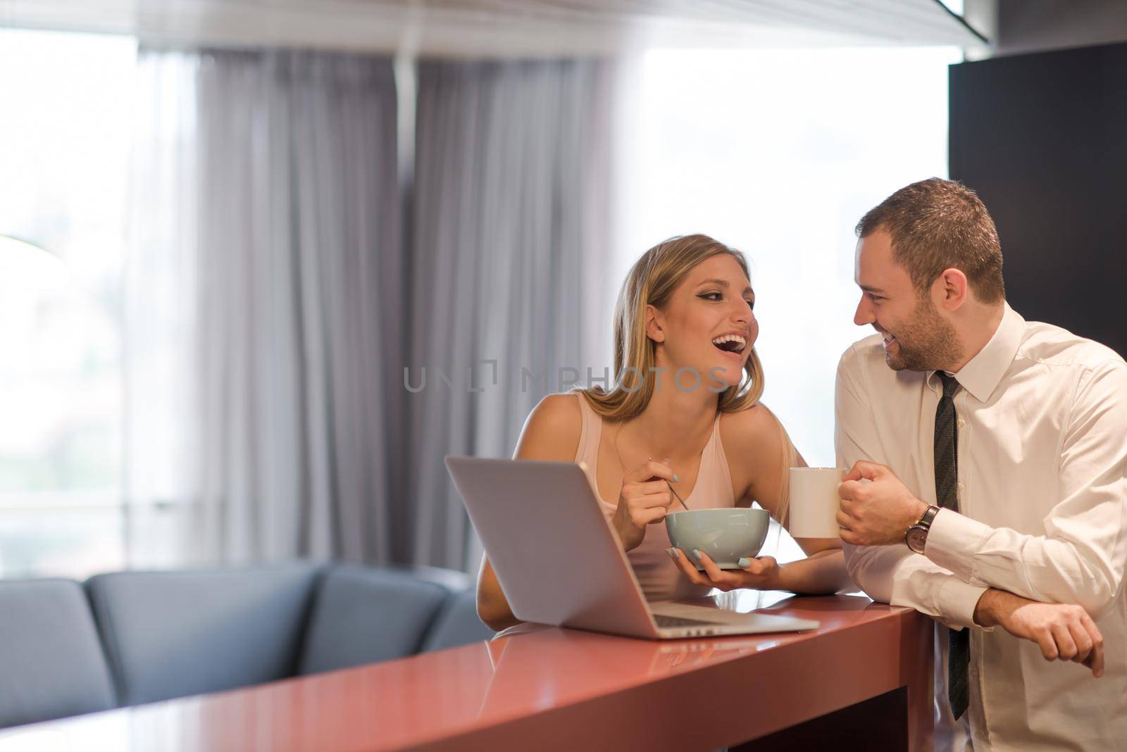 A young couple is preparing for the job and using a laptop. The man drinks coffee while the woman eats breakfast at luxury home together, looking at screen, smiling.