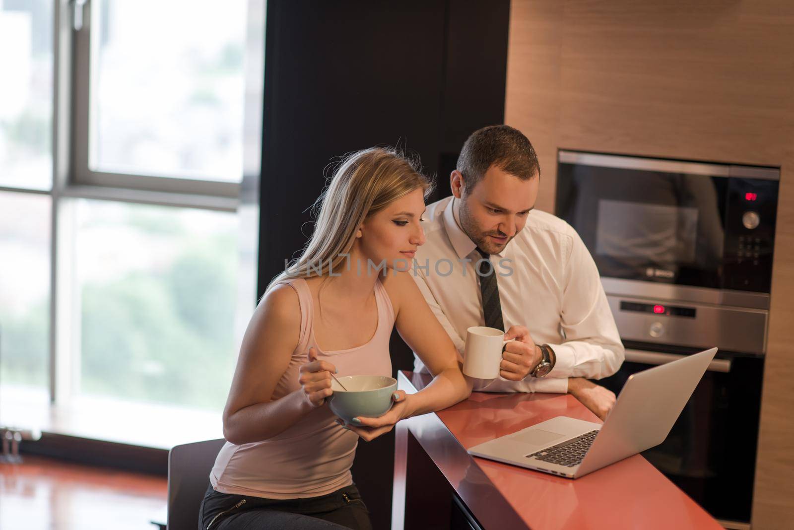 A young couple is preparing for the job and using a laptop. The man drinks coffee while the woman eats breakfast at luxury home together, looking at screen, smiling.