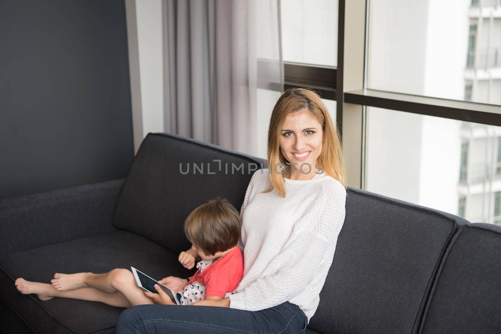 Beautiful young mother and her cute little daughter are using a tablet and smiling, sitting on sofa at home
