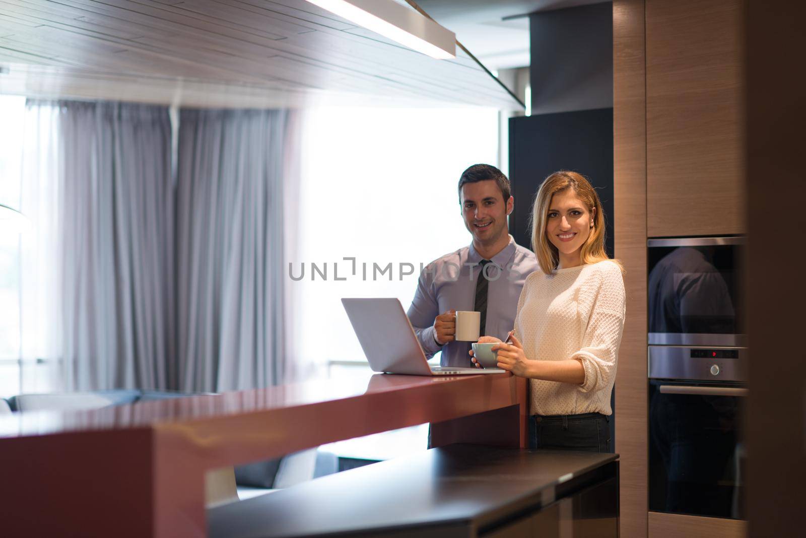 A young couple is preparing for the job and using a laptop. The man drinks coffee while the woman eats breakfast at luxury home together, looking at screen, smiling.