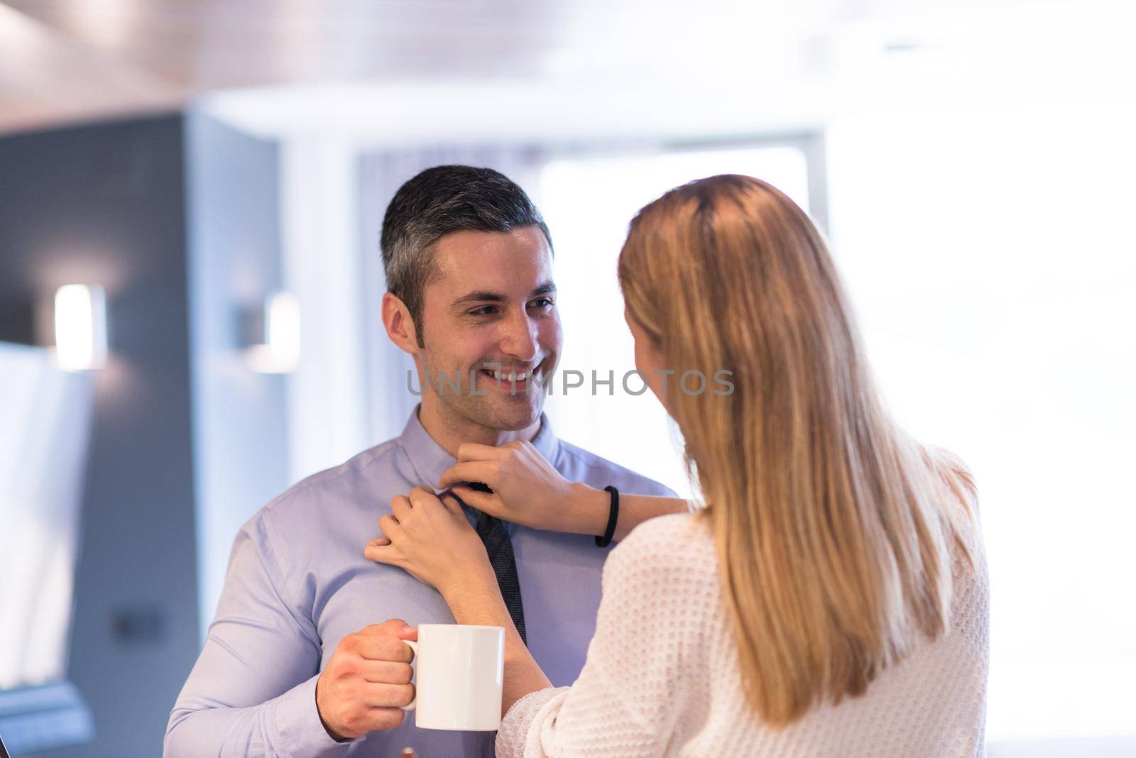 A young couple is preparing for the job and using a laptop. The man drinks coffee while the woman eats breakfast at luxury home together, looking at screen, smiling.