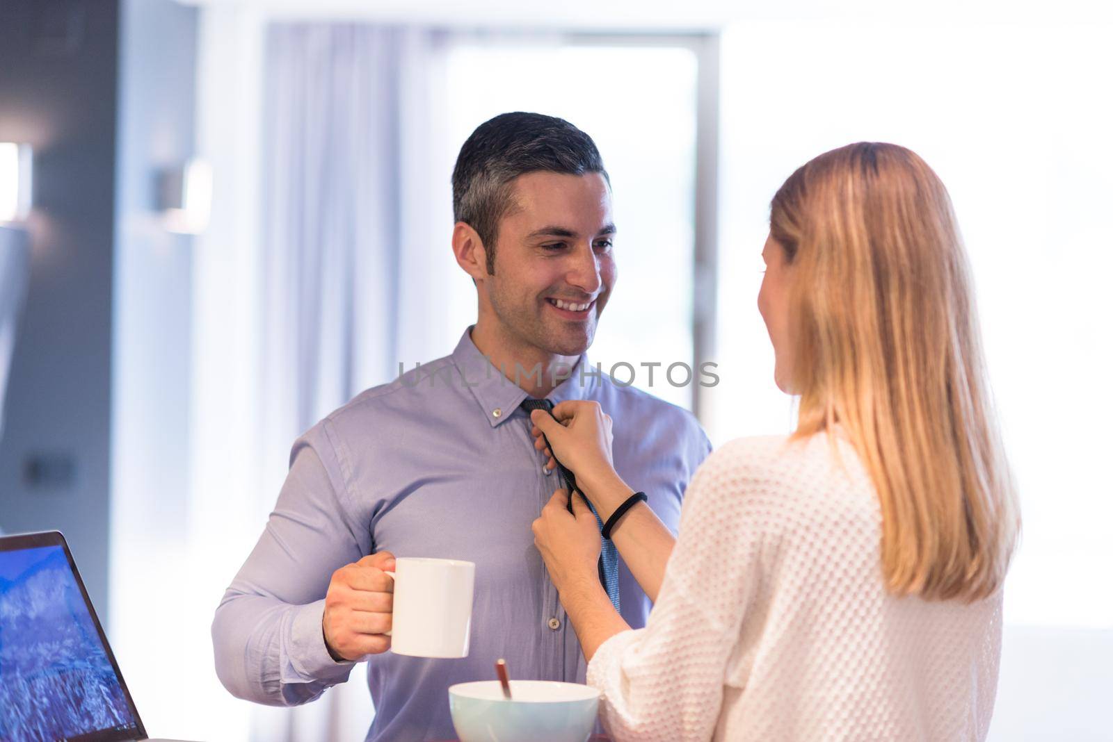 A young couple is preparing for the job and using a laptop. The man drinks coffee while the woman eats breakfast at luxury home together, looking at screen, smiling.