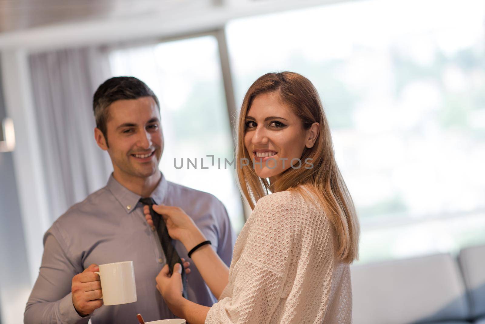 A young couple is preparing for the job and using a laptop. The man drinks coffee while the woman eats breakfast at luxury home together, looking at screen, smiling.