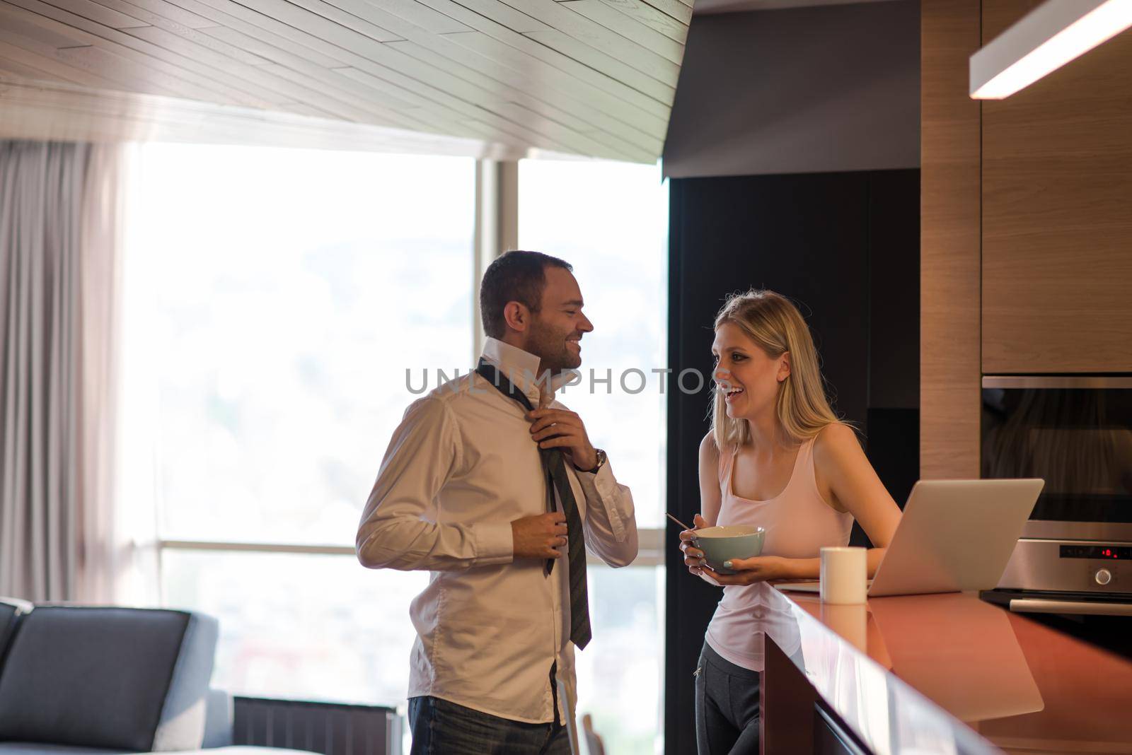 A young couple is preparing for the job and using a laptop. The man drinks coffee while the woman eats breakfast at luxury home together, looking at screen, smiling.