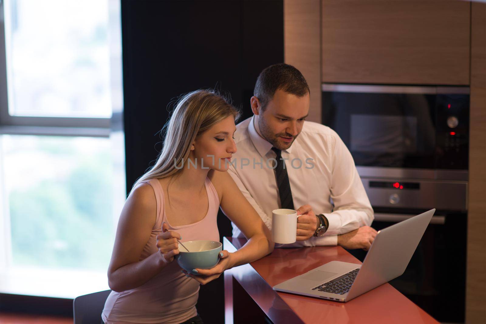 A young couple is preparing for the job and using a laptop. The man drinks coffee while the woman eats breakfast at luxury home together, looking at screen, smiling.