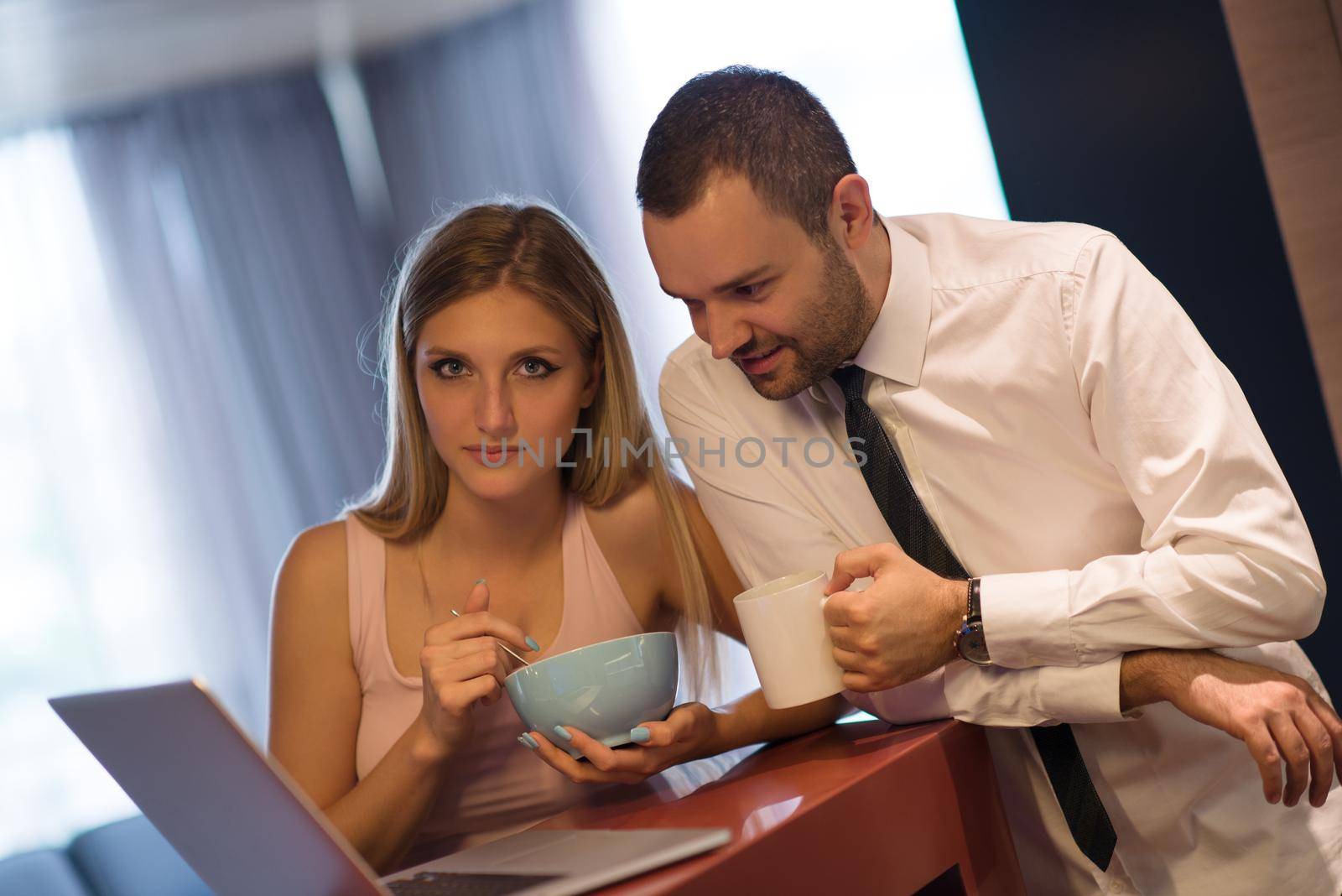 A young couple is preparing for the job and using a laptop. The man drinks coffee while the woman eats breakfast at luxury home together, looking at screen, smiling.