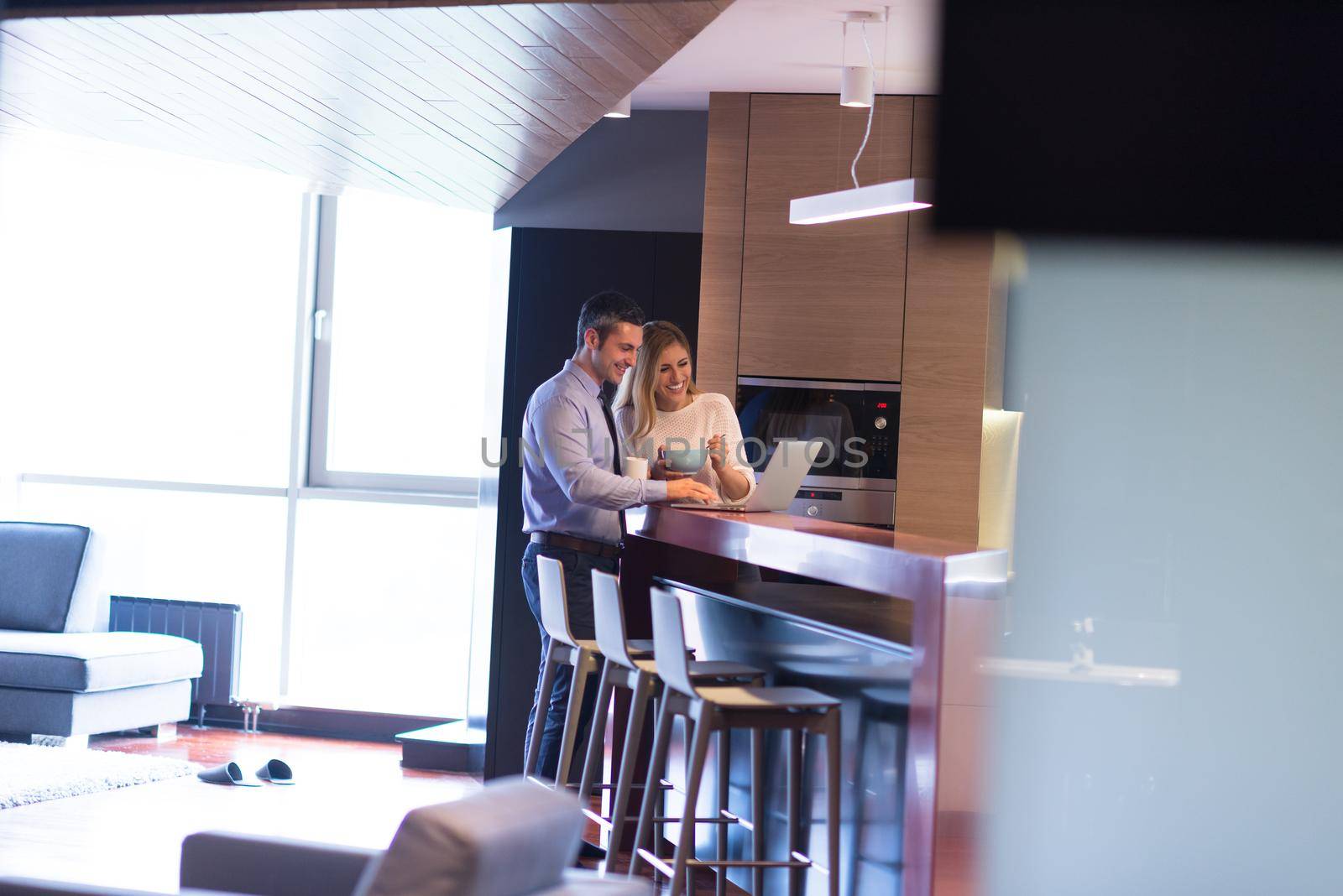 A young couple is preparing for the job and using a laptop. The man drinks coffee while the woman eats breakfast at luxury home together, looking at screen, smiling.