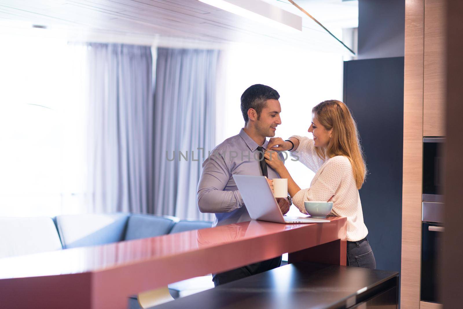 A young couple is preparing for the job and using a laptop. The man drinks coffee while the woman eats breakfast at luxury home together, looking at screen, smiling.