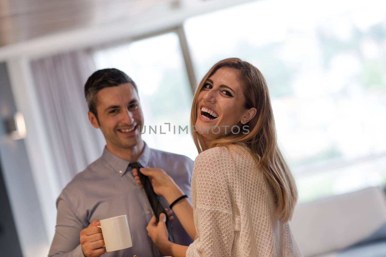 A young couple is preparing for the job and using a laptop. The man drinks coffee while the woman eats breakfast at luxury home together, looking at screen, smiling.