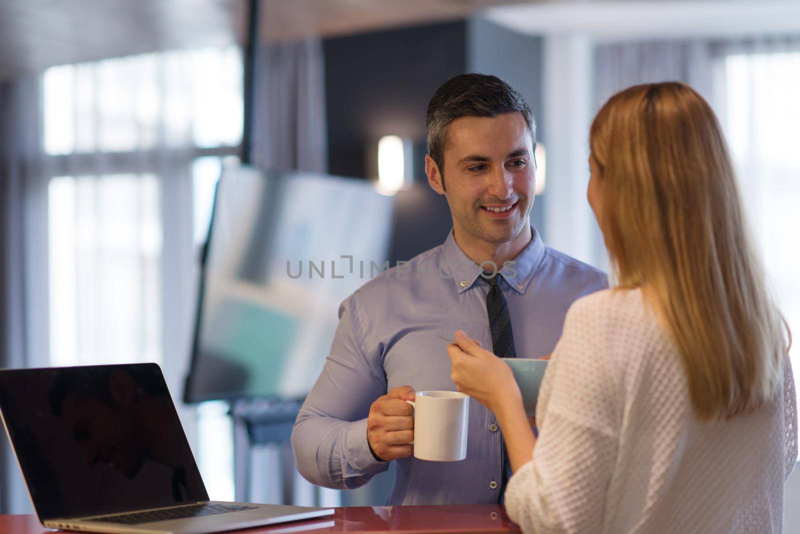 A young couple is preparing for the job and using a laptop. The man drinks coffee while the woman eats breakfast at luxury home together, looking at screen, smiling.