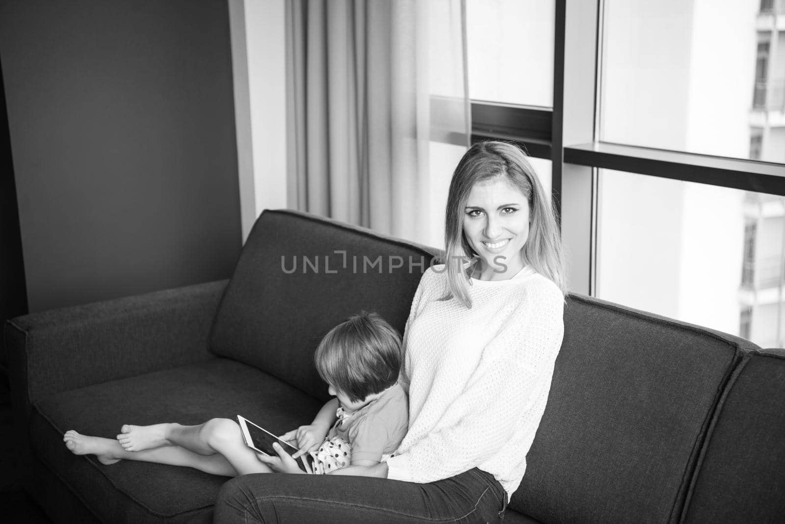 Beautiful young mother and her cute little daughter are using a tablet and smiling, sitting on sofa at home