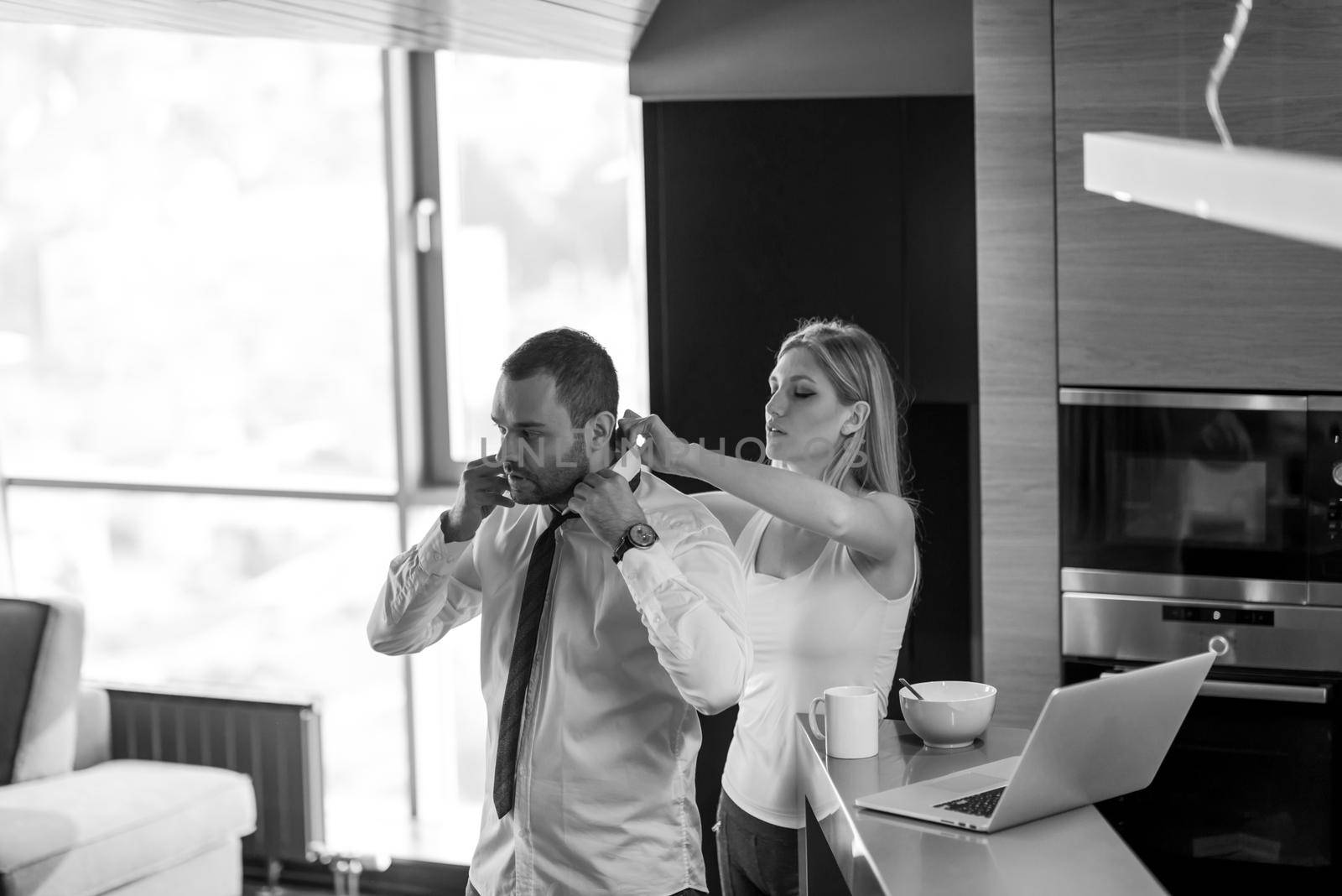 A young couple is preparing for the job and using a laptop. The man drinks coffee while the woman eats breakfast at luxury home together, looking at screen, smiling.