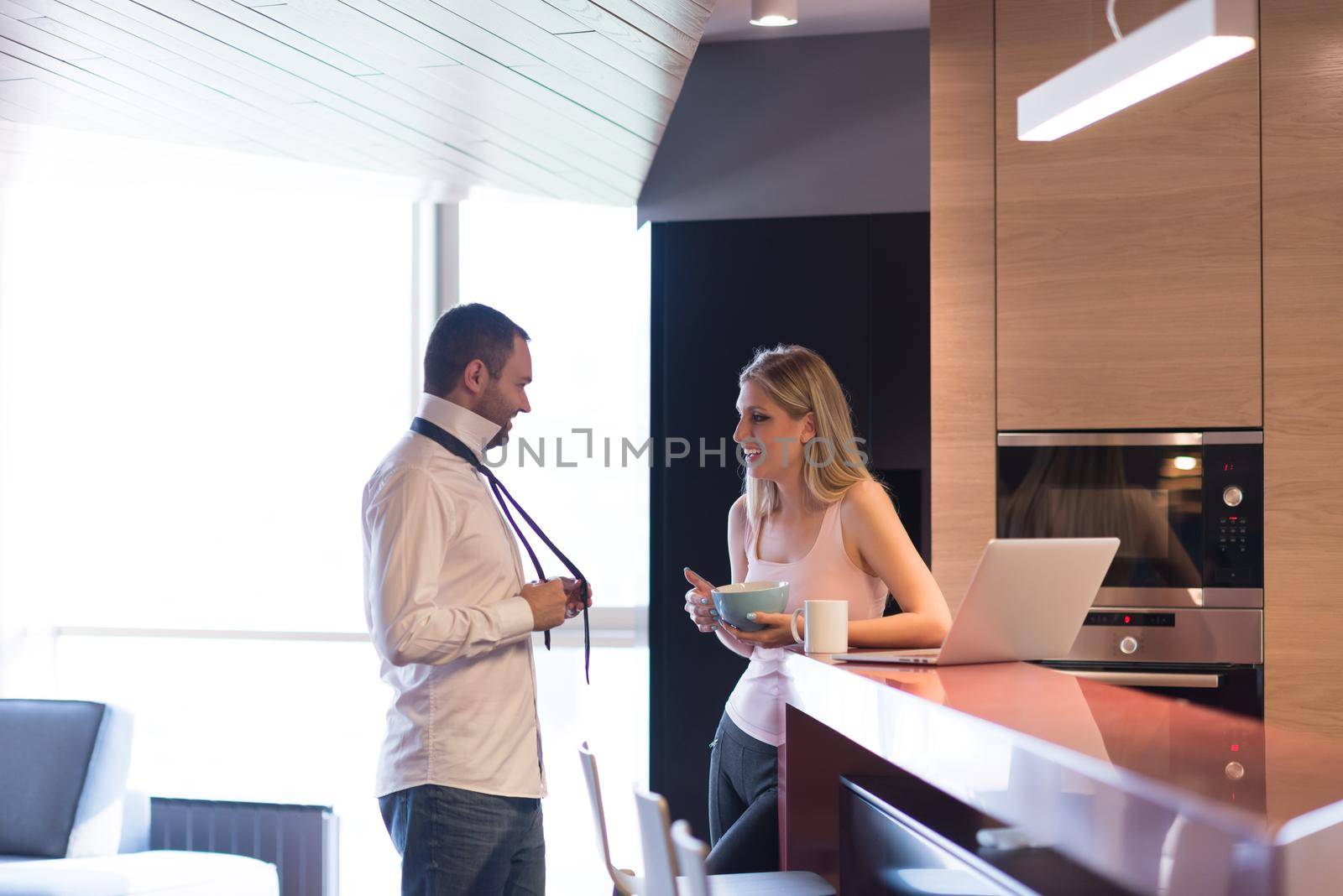 A young couple is preparing for the job and using a laptop. The man drinks coffee while the woman eats breakfast at luxury home together, looking at screen, smiling.