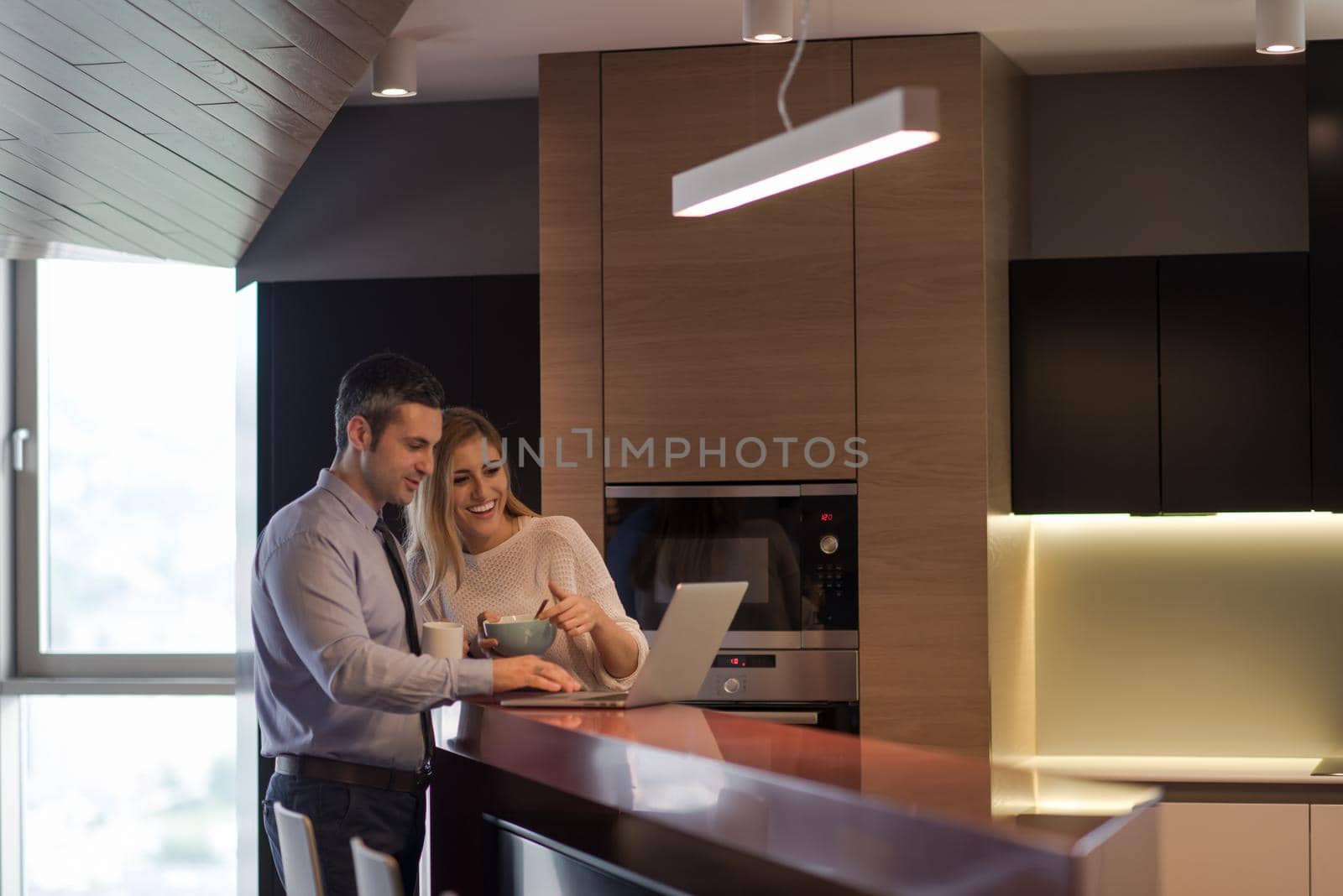 A young couple is preparing for the job and using a laptop. The man drinks coffee while the woman eats breakfast at luxury home together, looking at screen, smiling.