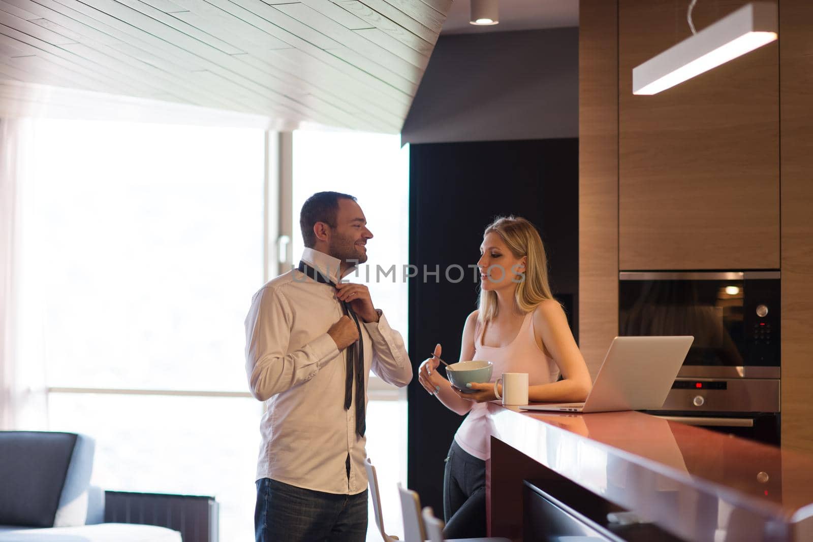 A young couple is preparing for the job and using a laptop. The man drinks coffee while the woman eats breakfast at luxury home together, looking at screen, smiling.