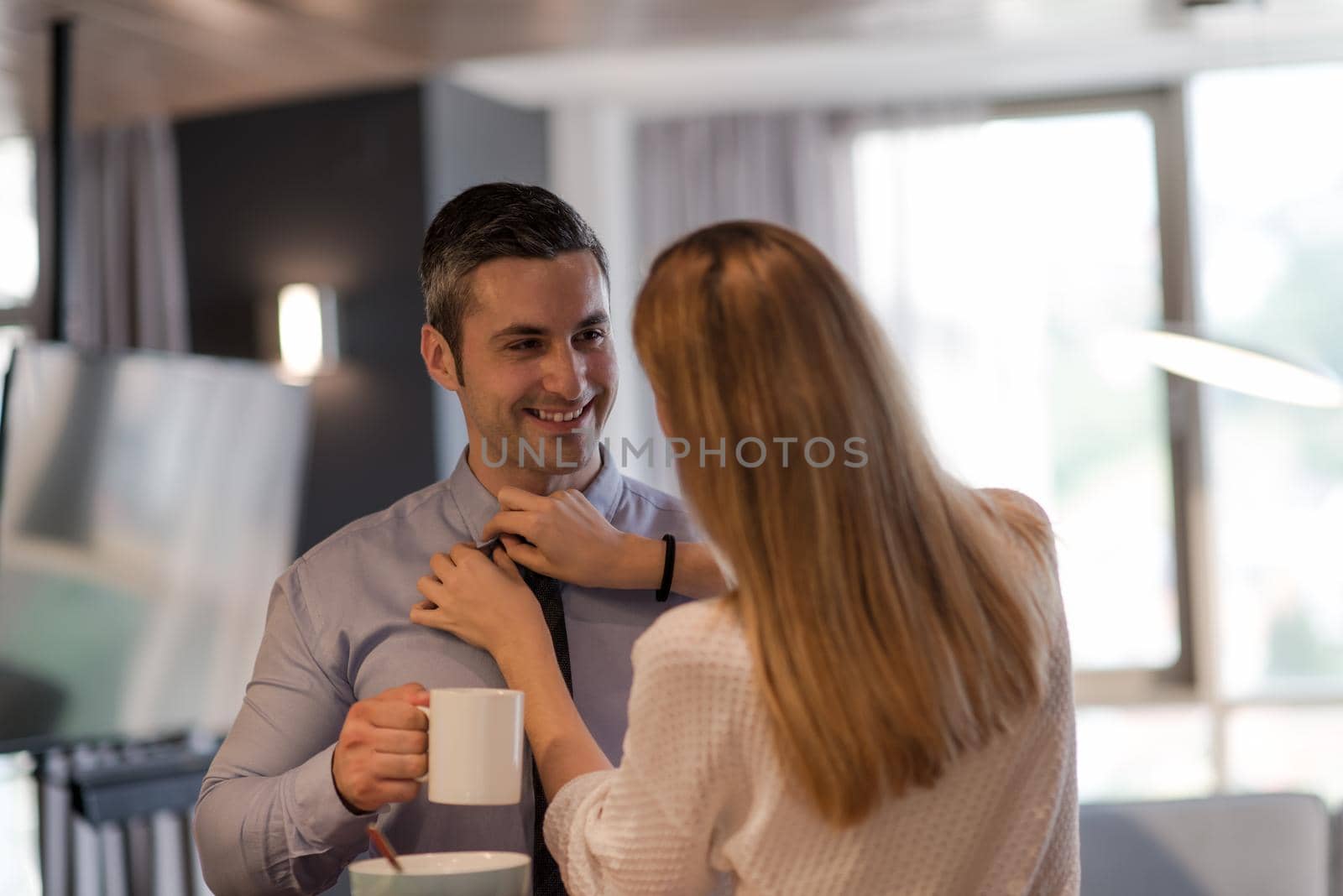 A young couple is preparing for the job and using a laptop. The man drinks coffee while the woman eats breakfast at luxury home together, looking at screen, smiling.