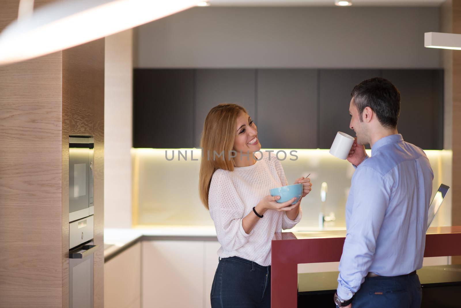 A young couple is preparing for the job and using a laptop. The man drinks coffee while the woman eats breakfast at luxury home together, looking at screen, smiling.