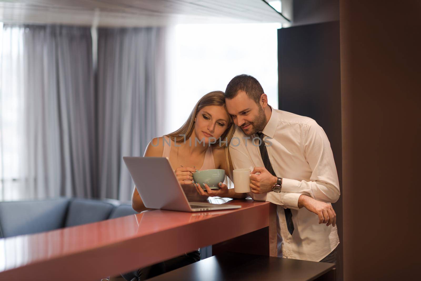 A young couple is preparing for the job and using a laptop. The man drinks coffee while the woman eats breakfast at luxury home together, looking at screen, smiling.