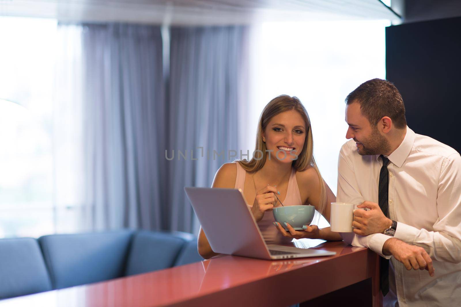 A young couple is preparing for the job and using a laptop. The man drinks coffee while the woman eats breakfast at luxury home together, looking at screen, smiling.