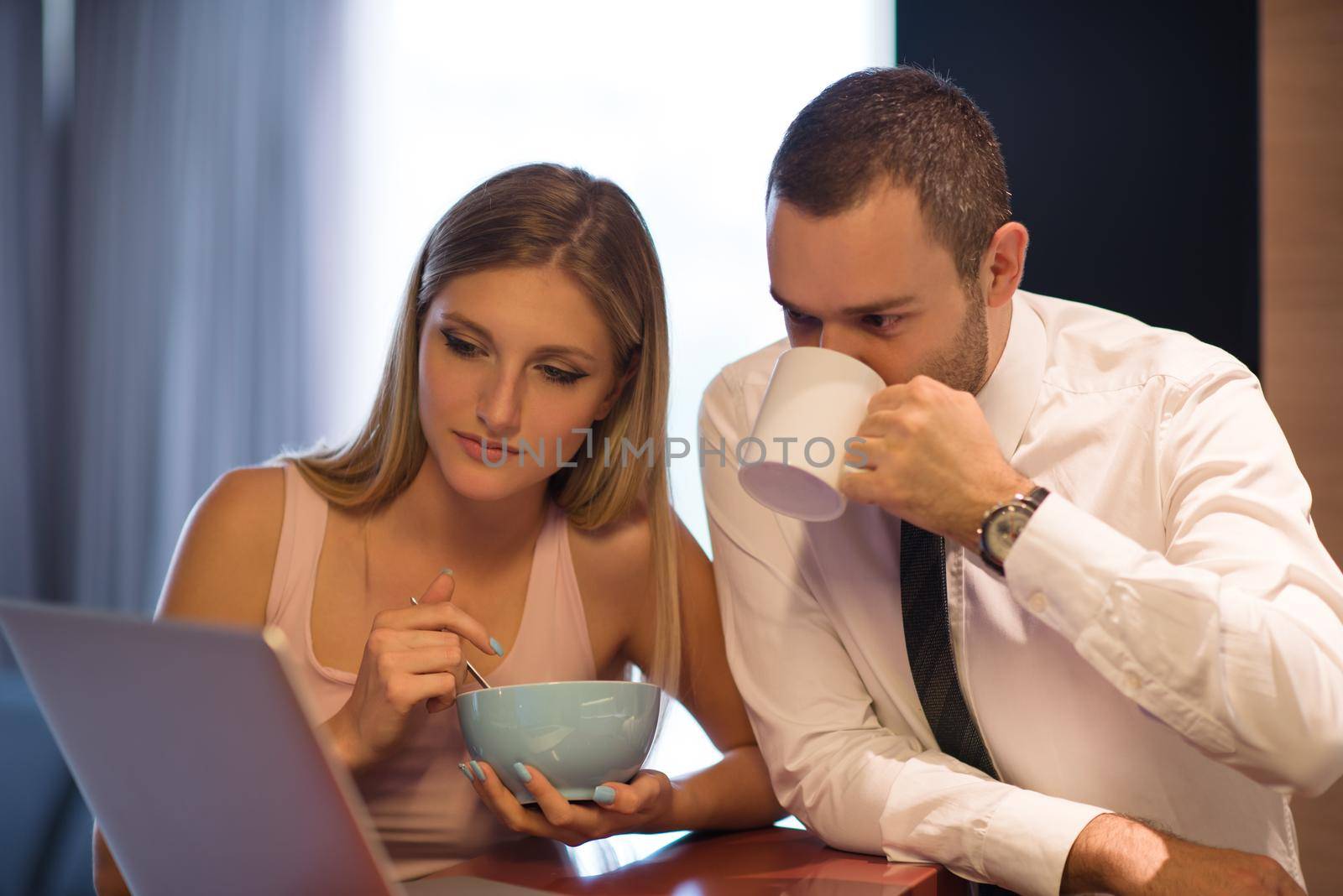 A young couple is preparing for the job and using a laptop. The man drinks coffee while the woman eats breakfast at luxury home together, looking at screen, smiling.