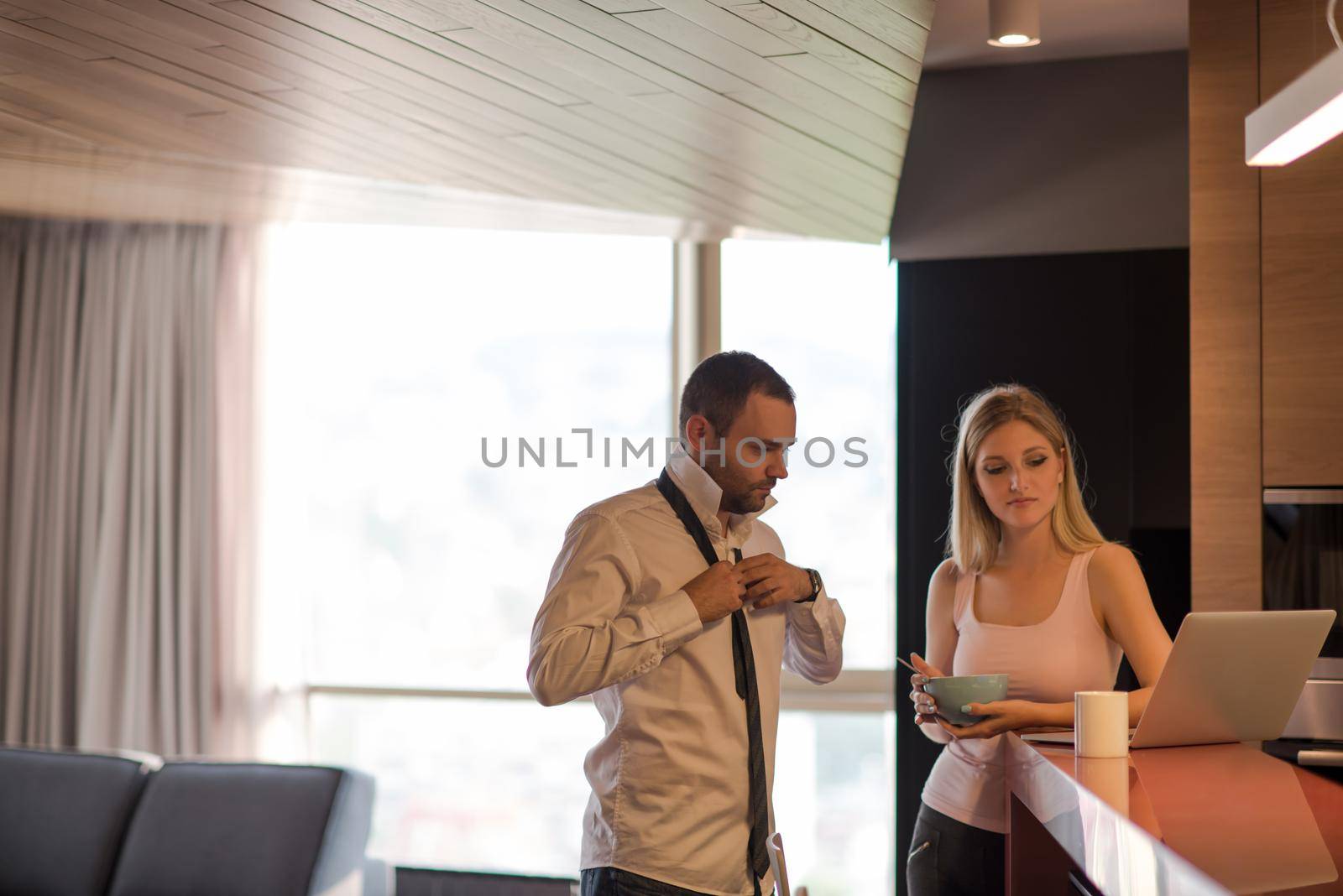 A young couple is preparing for the job and using a laptop. The man drinks coffee while the woman eats breakfast at luxury home together, looking at screen, smiling.