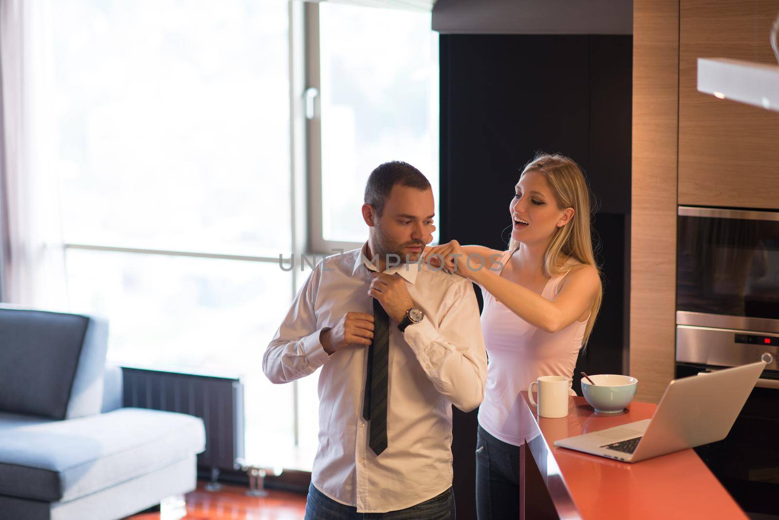 A young couple is preparing for the job and using a laptop. The man drinks coffee while the woman eats breakfast at luxury home together, looking at screen, smiling.