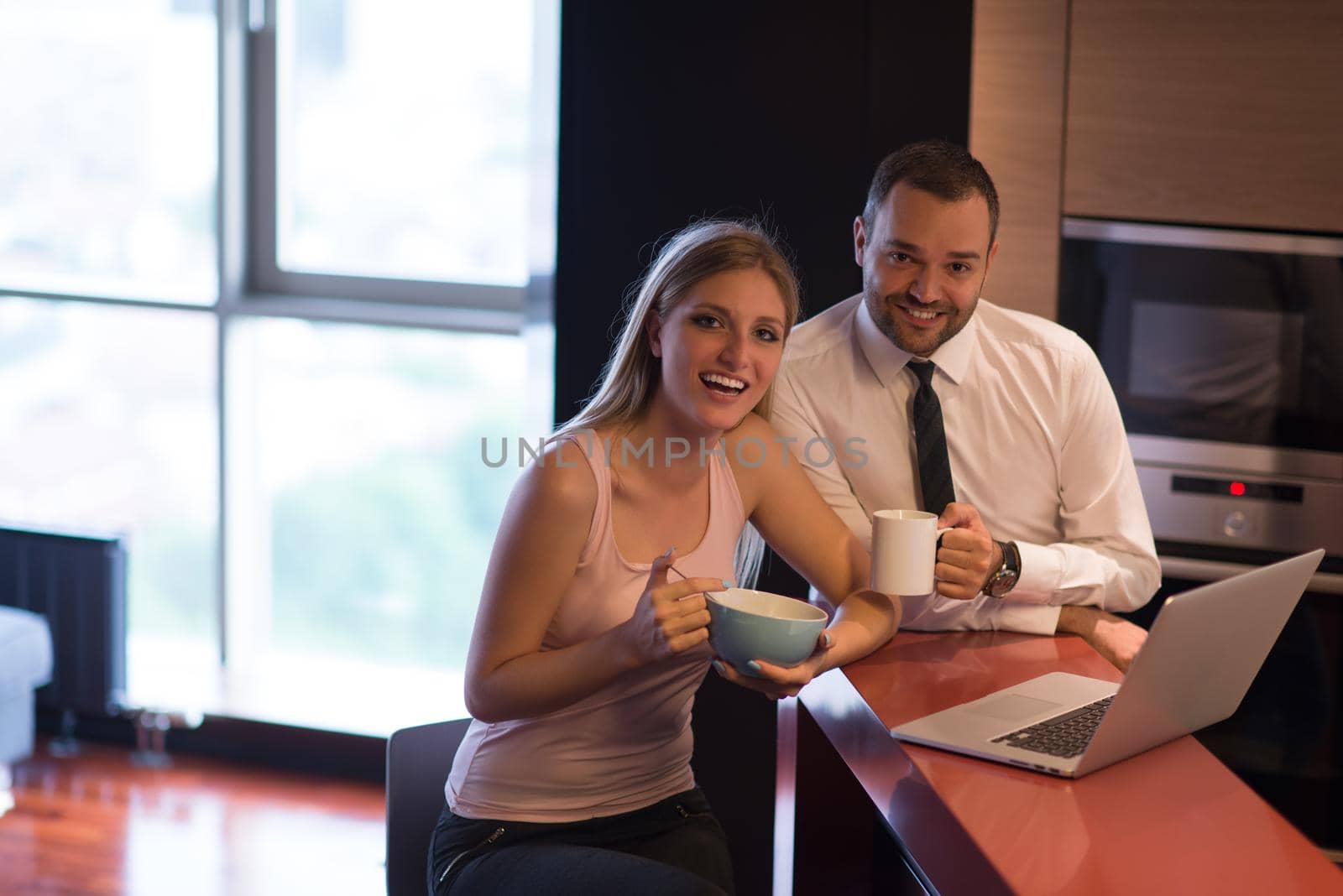 A young couple is preparing for the job and using a laptop. The man drinks coffee while the woman eats breakfast at luxury home together, looking at screen, smiling.