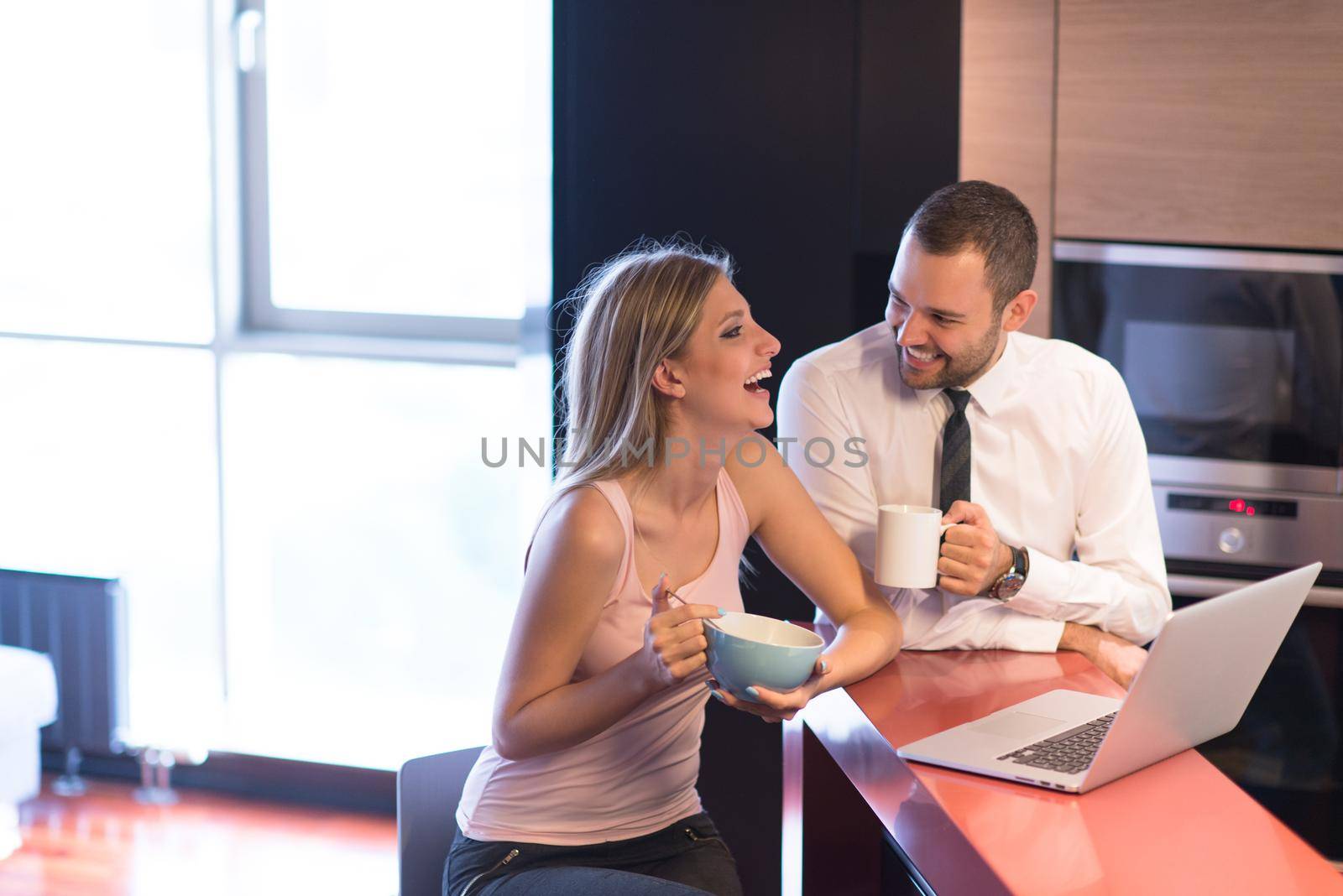 A young couple is preparing for the job and using a laptop. The man drinks coffee while the woman eats breakfast at luxury home together, looking at screen, smiling.