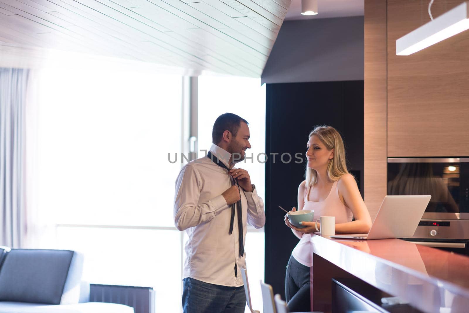 A young couple is preparing for the job and using a laptop. The man drinks coffee while the woman eats breakfast at luxury home together, looking at screen, smiling.