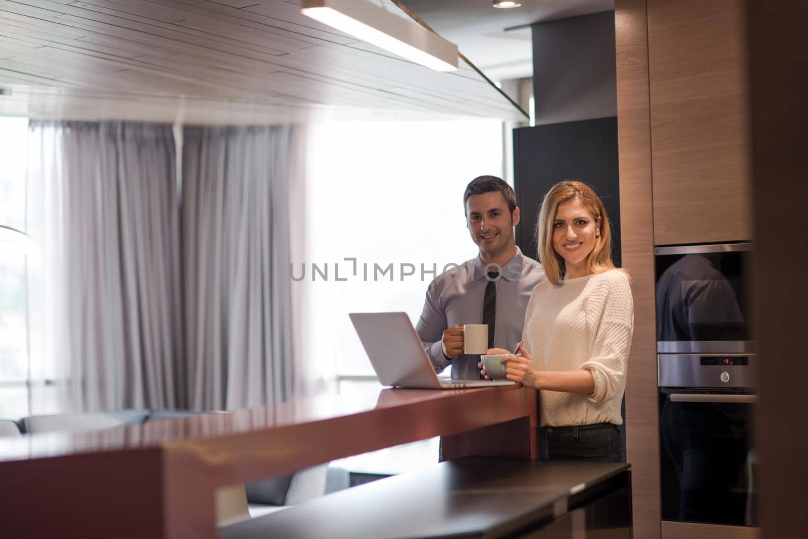 A young couple is preparing for the job and using a laptop. The man drinks coffee while the woman eats breakfast at luxury home together, looking at screen, smiling.