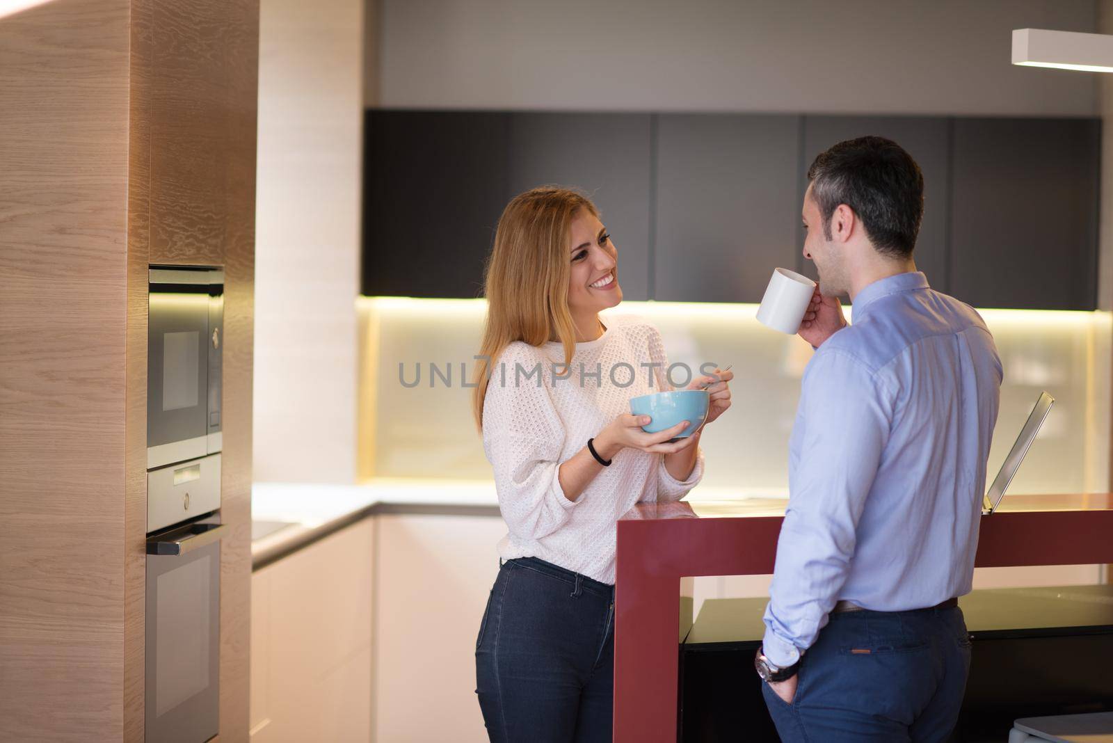 A young couple is preparing for the job and using a laptop. The man drinks coffee while the woman eats breakfast at luxury home together, looking at screen, smiling.