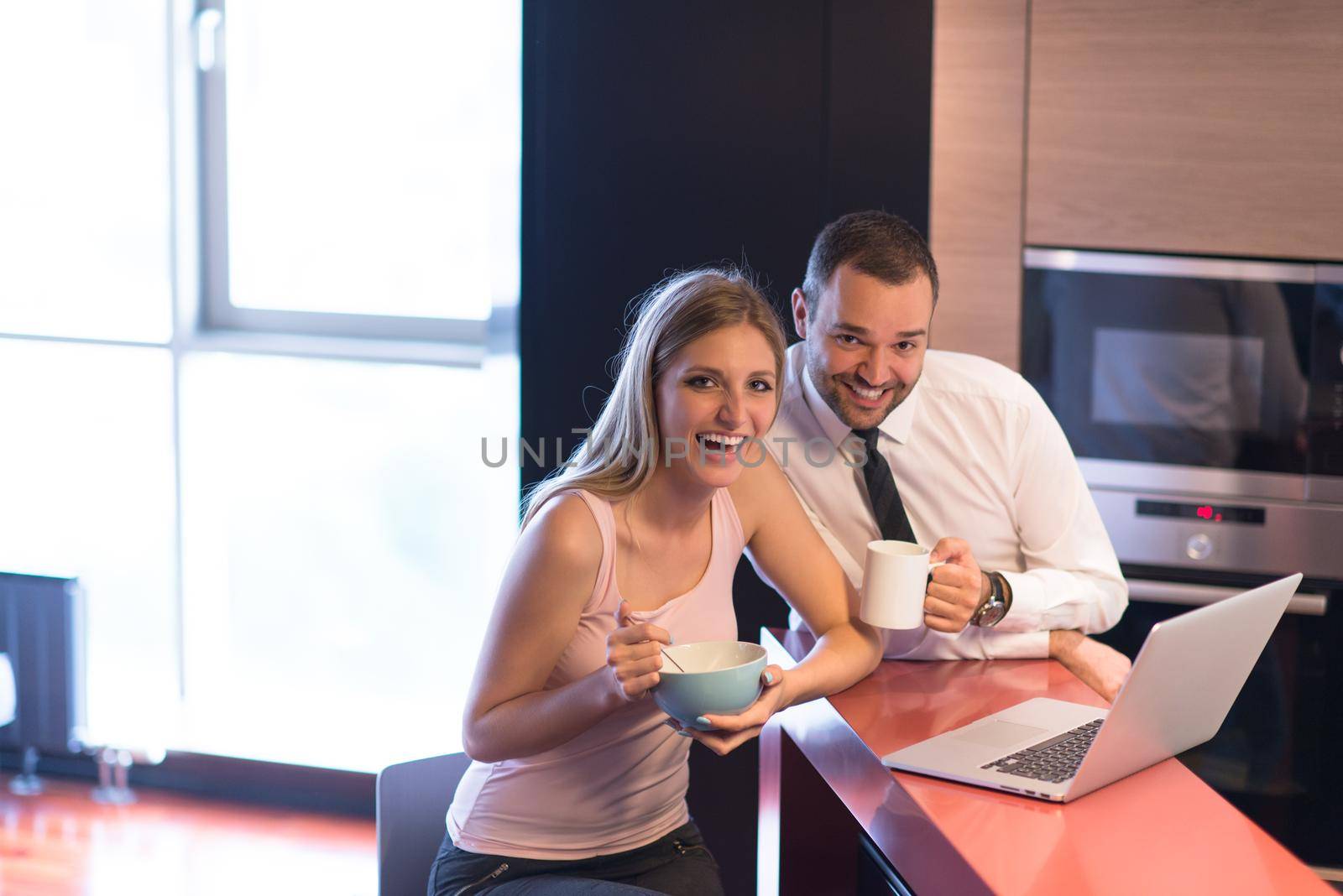 A young couple is preparing for the job and using a laptop. The man drinks coffee while the woman eats breakfast at luxury home together, looking at screen, smiling.