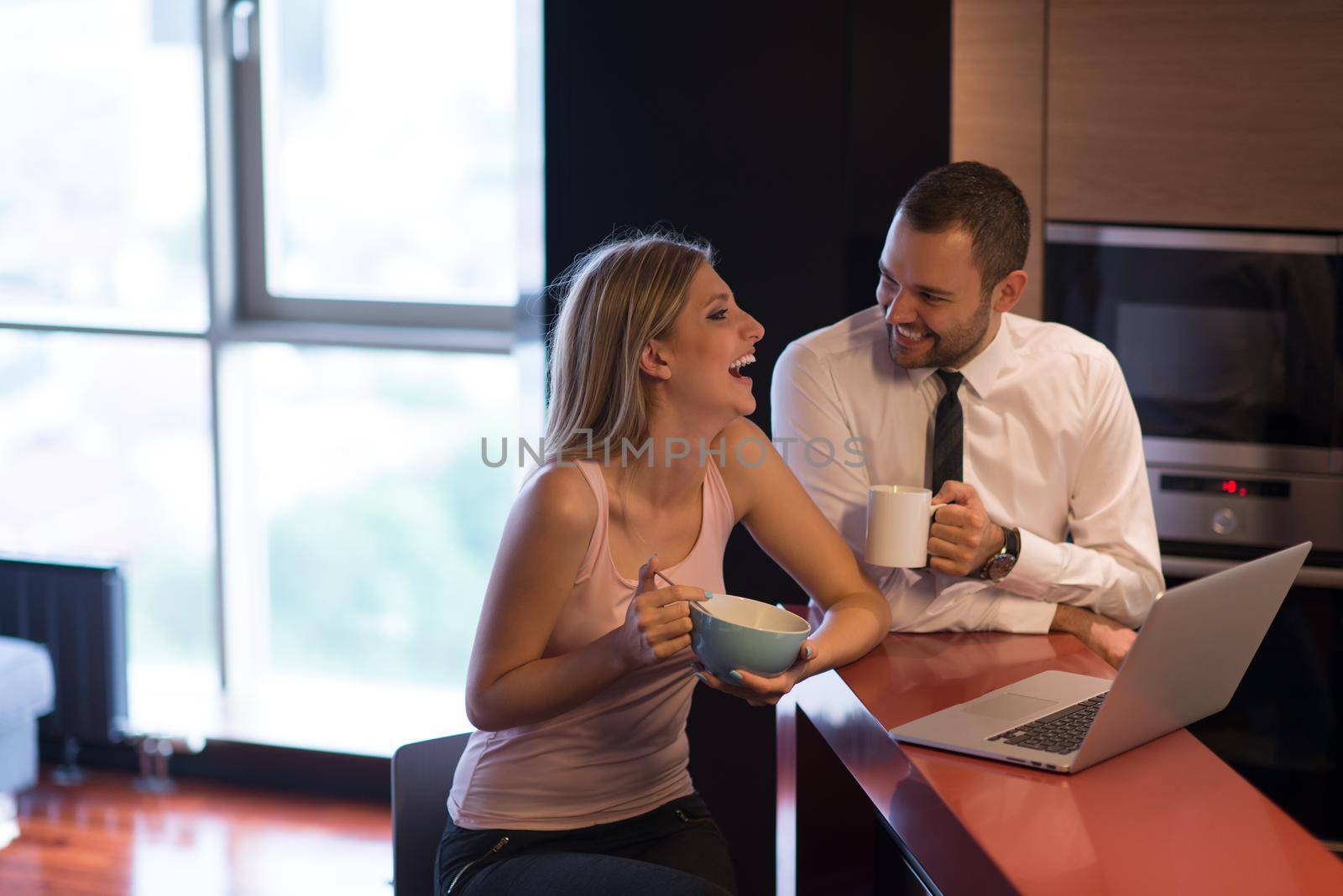 A young couple is preparing for the job and using a laptop. The man drinks coffee while the woman eats breakfast at luxury home together, looking at screen, smiling.