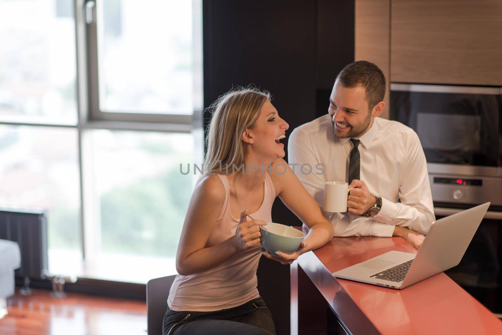 A young couple is preparing for the job and using a laptop. The man drinks coffee while the woman eats breakfast at luxury home together, looking at screen, smiling.