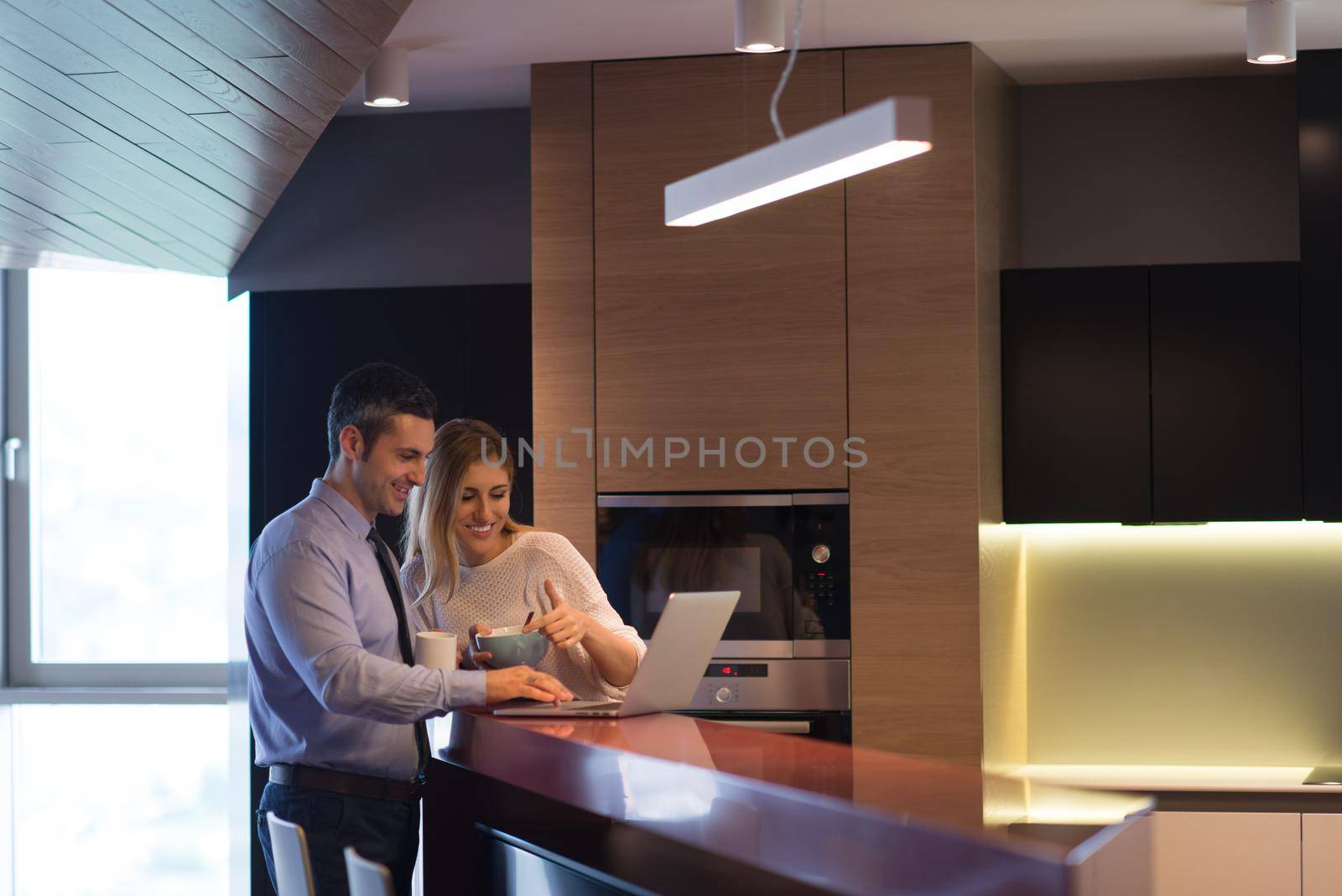 A young couple is preparing for the job and using a laptop. The man drinks coffee while the woman eats breakfast at luxury home together, looking at screen, smiling.