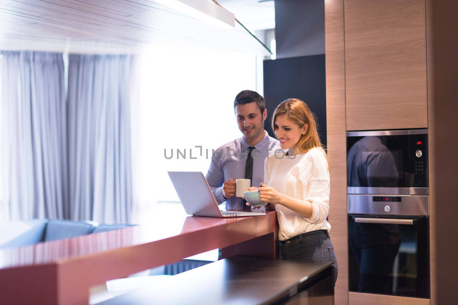 A young couple is preparing for the job and using a laptop. The man drinks coffee while the woman eats breakfast at luxury home together, looking at screen, smiling.