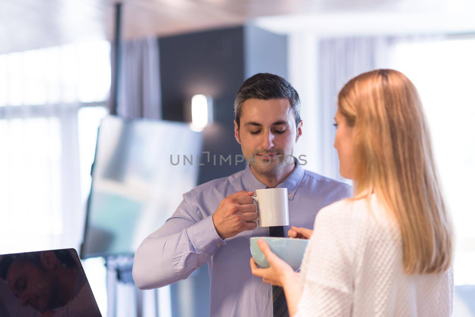 A young couple is preparing for the job and using a laptop. The man drinks coffee while the woman eats breakfast at luxury home together, looking at screen, smiling.