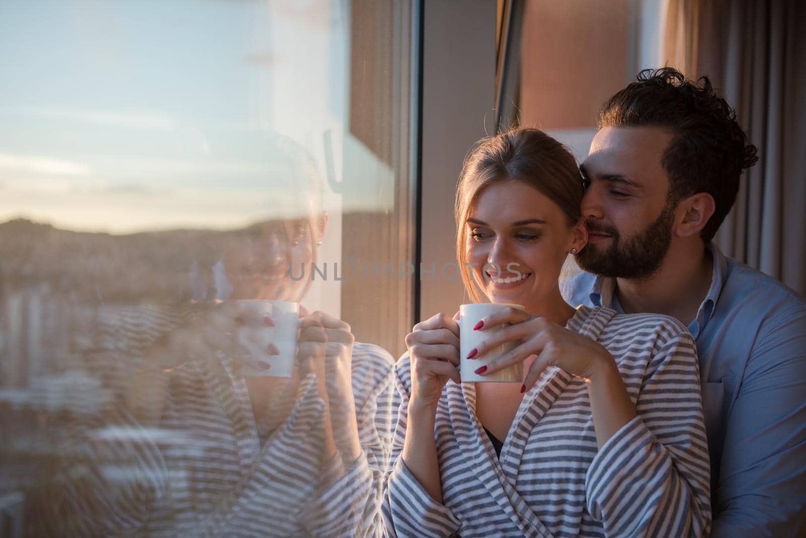 romantic happy young couple enjoying evening coffee and beautiful sunset landscape of the city while standing by the window