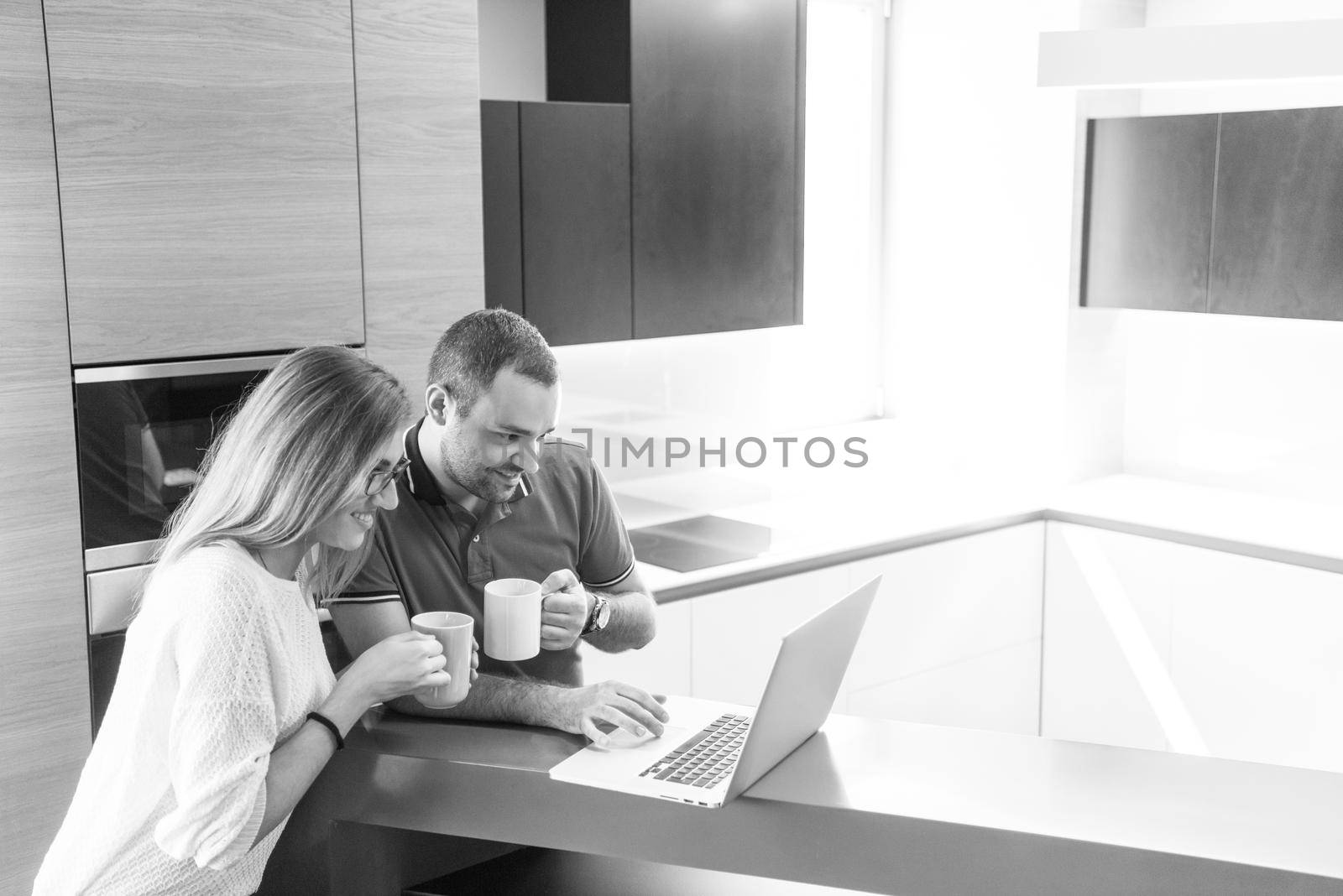 Young couple drinking coffee and using laptop computer at luxury home together, looking at screen, smiling.