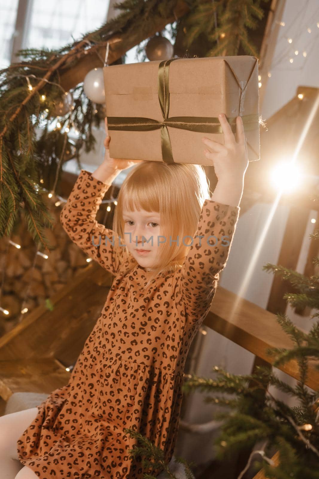 A little blonde girl is sitting on a wooden staircase in a Scandinavian interior decorated in a New Year's style. A child holds a Christmas gift in a craft package.