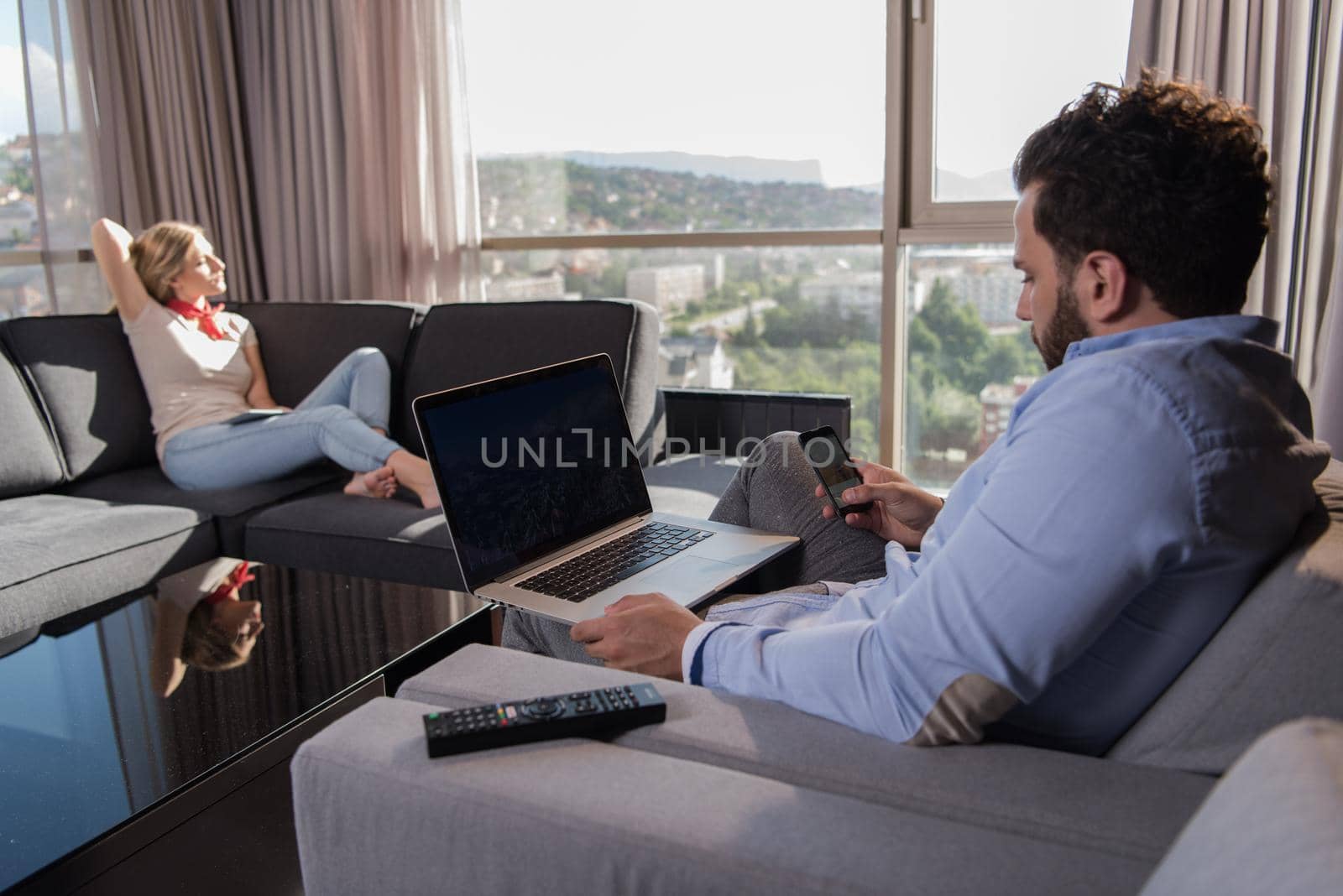 Young couple relaxing at home using tablet and laptop computers reading near the window on the sofa couch.