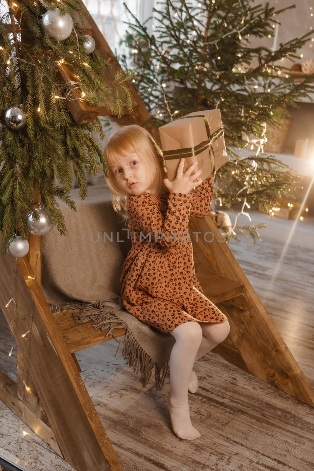 A little blonde girl is sitting on a wooden staircase in a Scandinavian interior decorated in a New Year's style. A child holds a Christmas gift in a craft package.