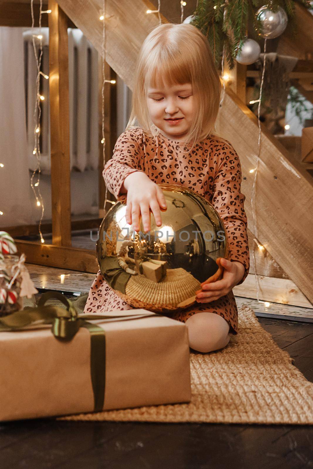 A little blonde girl is sitting next to a wooden staircase in a Scandinavian interior decorated in a New Year's style. A child holds a golden Christmas ball.