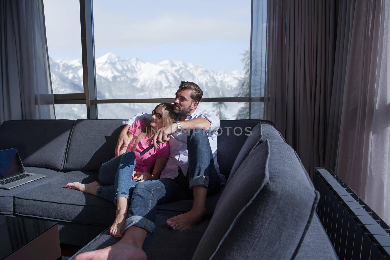 Young couple sitting on sofa near the window watching television together at luxury home