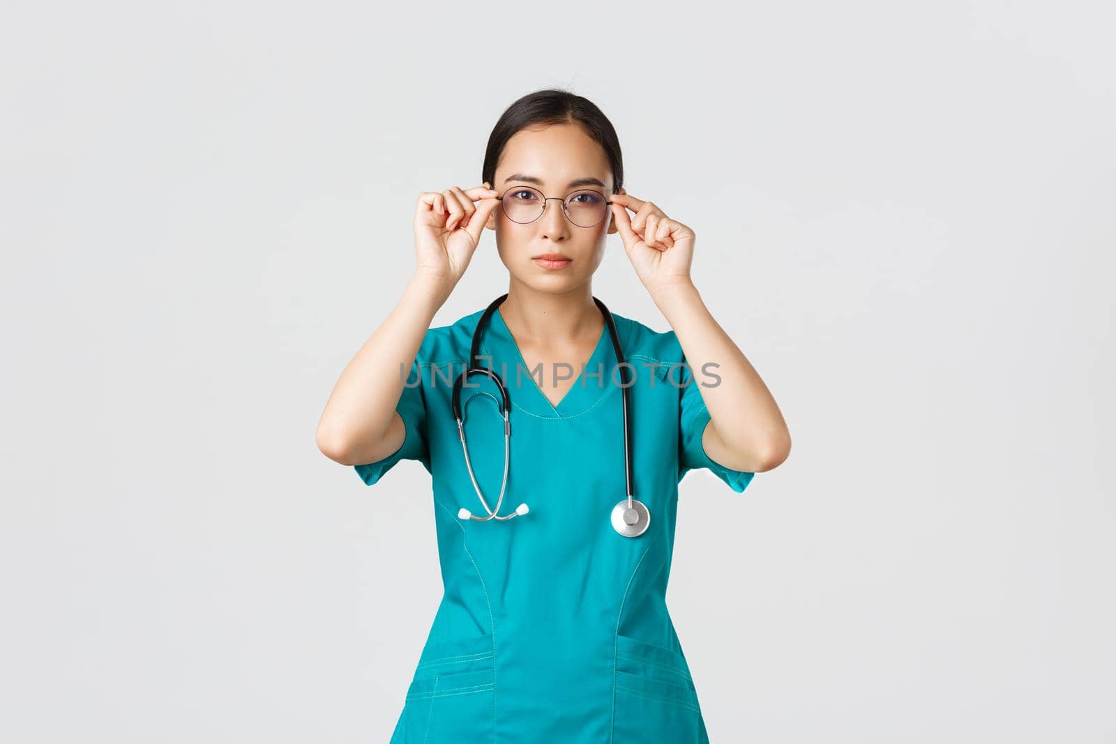 Covid-19, healthcare workers, pandemic concept. Confident serious-looking and determined asian female doctor, nurse in scrubs put on glasses, ready for shift in emergency room, white background by Benzoix