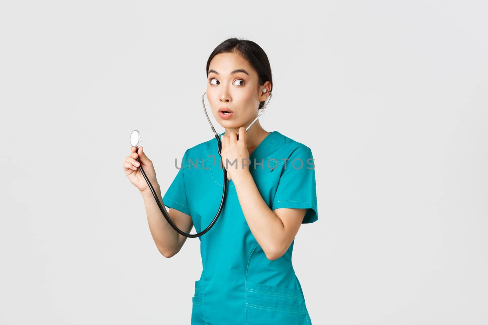 Covid-19, healthcare workers and preventing virus concept. Intrigued asian female doctor, physician in scrubs, looking away thoughtful, listening to patient lungs with stethoscope, white background.