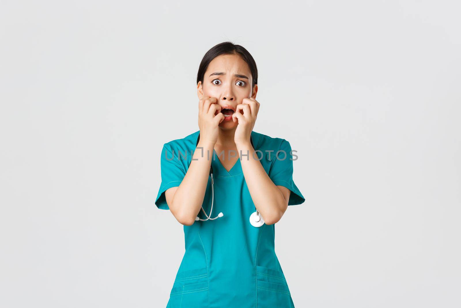 Covid-19, healthcare workers and preventing virus concept. Scared insecure asian female doctor, nurse in scrubs starts to panic from fear, biting fingernails and looking horrified, white background.