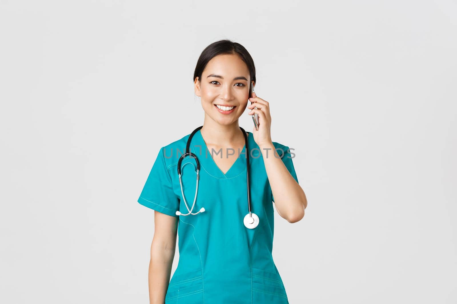 Covid-19, healthcare workers and preventing virus concept. Smiling happy asian female doctor, young intern in scrubs talking on phone, looking at camera hopeful, white background.